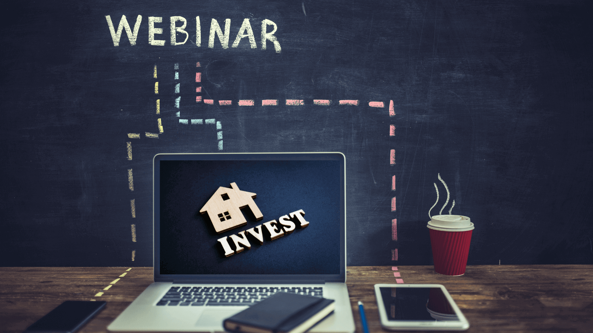Laptop mit dem Wort 'INVEST' und einem Haus-Symbol auf dem Bildschirm, daneben ein Notizbuch und eine Kaffeetasse.